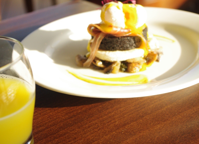 Plated stacked breakfast dish with poached egg, sauce, and dark bread on a white plate, glass of orange juice beside it