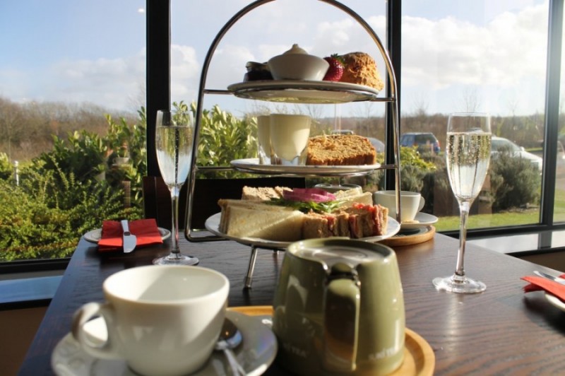 Three-tier stand with sandwiches, scones and cakes on a table with teapot, cups and champagne glasses by a window overlooking greenery