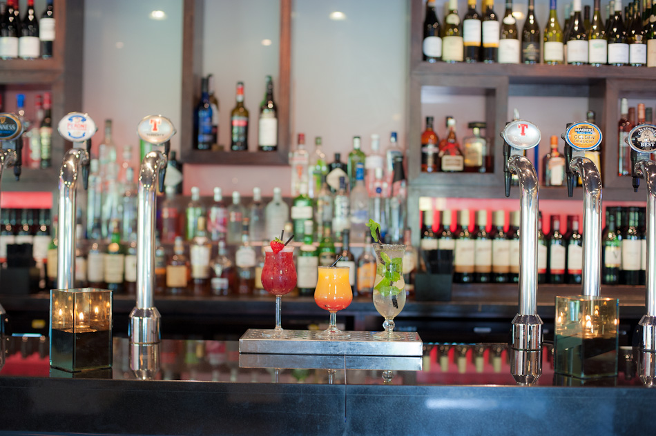 Bar counter with beer taps and three colorful cocktails, shelves of assorted bottles in background, warm ambient lighting