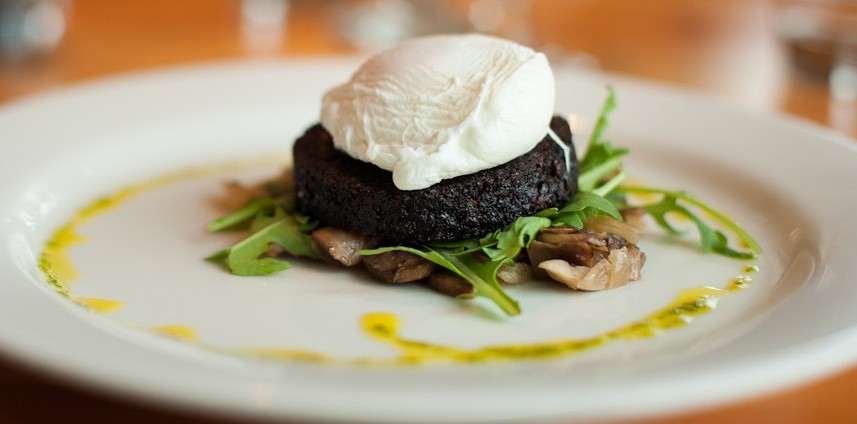 A dark round food patty topped with a poached egg, served on arugula and mushrooms with yellow sauce drizzle on white plate