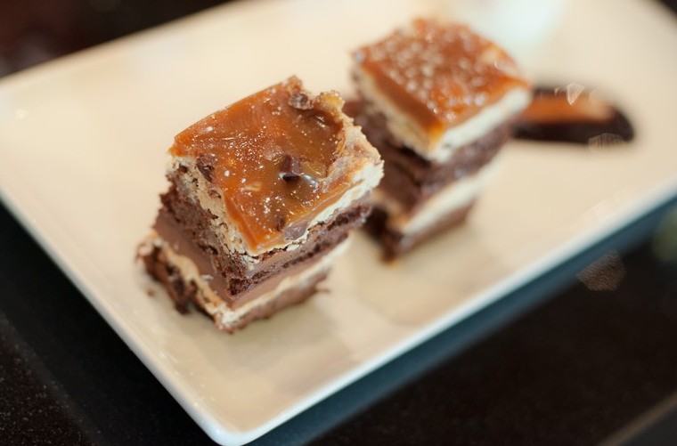 Two layered dessert bars with caramel topping and chocolate base on a white rectangular plate, warm brown and amber tones